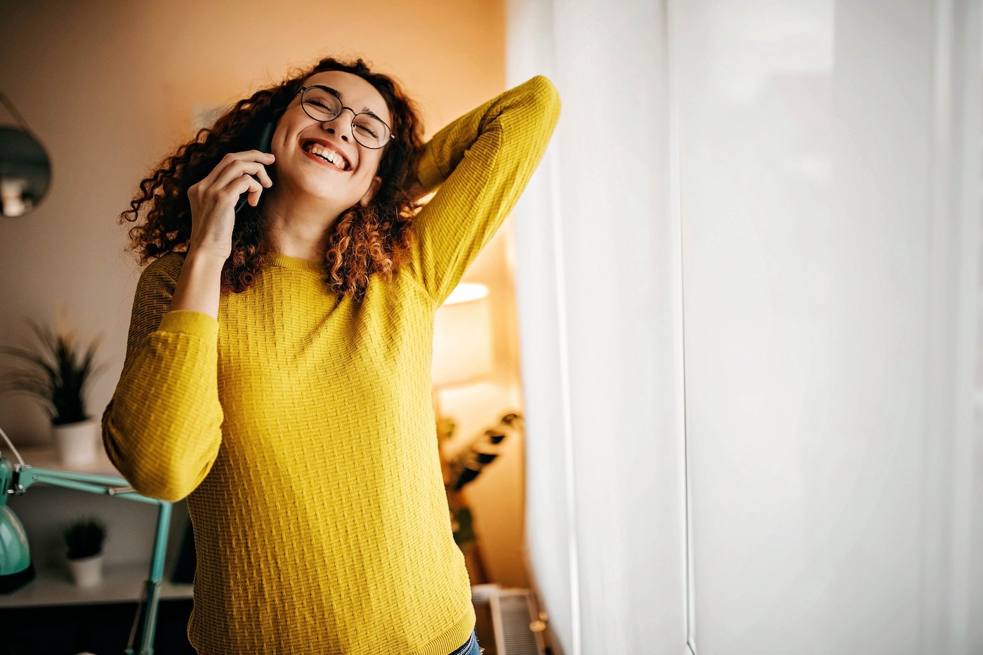 Woman in yellow sweater stretching and smiling while talking on her mobile phone, enjoying easy essential connections