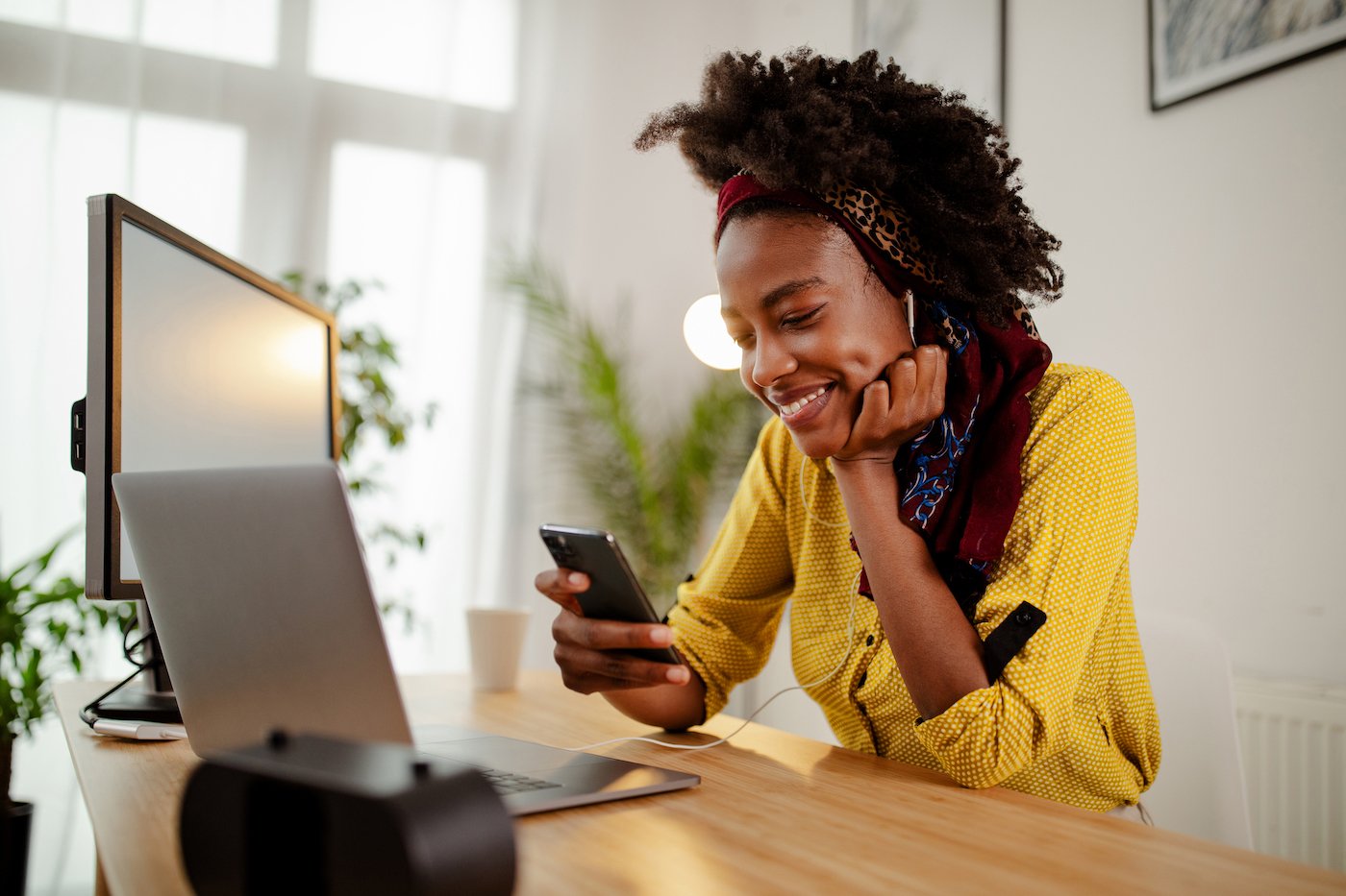 Woman wearing yellow looking at her phone and smiling next to laptop